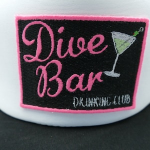 May include: White and black trucker hat with a pink embroidered patch that says "Dive Bar Drinking Club" and features a martini glass with a green olive.