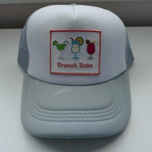 May include: A white and light blue trucker hat with a patch that says "Brunch Babe" and features an embroidered design of three cocktails.