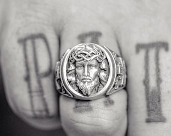 May include: A silver ring with a detailed design of a man's face with a crown of thorns. The ring is worn on a finger with the word "FAITH" tattooed on it.