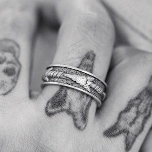 Op de afbeelding: Een zilveren bandring met een getextureerd oppervlak en een ontwerp van een gestileerde pijl. De ring wordt gedragen op de wijsvinger van een hand met verschillende tatoeages.