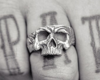 May include: A silver skull ring with a detailed design. The ring is worn on the middle finger of a hand with the letters "PIT" tattooed on the fingers.