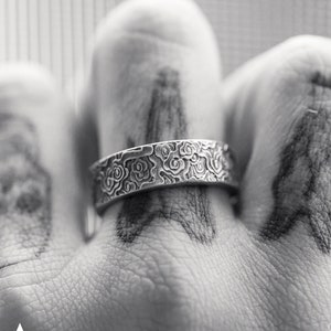 Può includere: Un anello d'argento con un motivo floreale è mostrato su una mano tatuata. L'anello presenta dettagliati disegni floreali. La parola "ACHILLES" è stampata in basso nell'immagine.