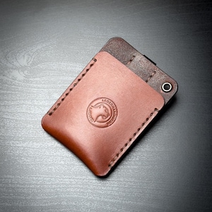May include: A rectangular leather card holder featuring a two-tone design with a light brown front pocket and a darker brown back section. The front pocket is stitched with thick thread and displays a circular embossed logo of a lion's head.