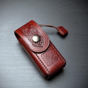 May include: A reddish-brown leather pouch featuring embossed Celtic knotwork patterns on the front flap and body. The rectangular case is secured with a single silver-toned metal snap button and includes a small leather pull tab attached to the side.