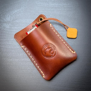 May include: A brown leather pouch with light-colored stitching, featuring a circular embossed logo of a lion's head and the text LEDERHANDWERK LION. A small red multi-tool is partially inserted, and a leather cord with a square tag is attached.
