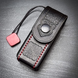 May include: A black leather case featuring embossed Celtic knot patterns on the front flap and body. The case is secured with a silver-toned metal snap button and accented with red stitching along the sides. A thin black cord with a red square tag.