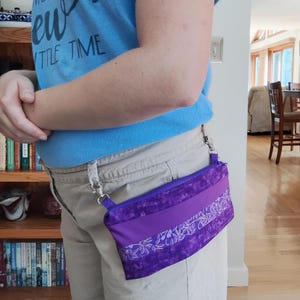 May include: A rectangular purple pouch with a zipper closure, featuring a solid purple center band flanked by two sections of purple fabric with a white floral pattern. The pouch is attached to belt loops with silver metal swivel lobster clasps.