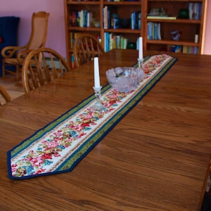 May include: A wooden dining table with a floral table runner, two candlesticks with white candles, and a glass bowl. The table is set with chairs and is in a room with a bookshelf in the background.