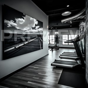 Può includere: Una fotografia in bianco e nero di una strada con un cielo nuvoloso è appesa al muro di una palestra. La palestra ha un pavimento in legno e tapis roulant.