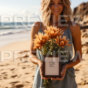 May include: A woman on a beach holds a bouquet of dried flowers and a framed print with the text "Wildly Made" and "Boho Vibes".