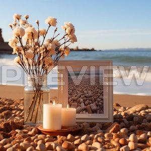May include: A white picture frame with a photo of a beach with smooth, round stones. Two white candles are in front of the frame, and a vase with white flowers is to the left. The scene is set on a beach with sand and stones.