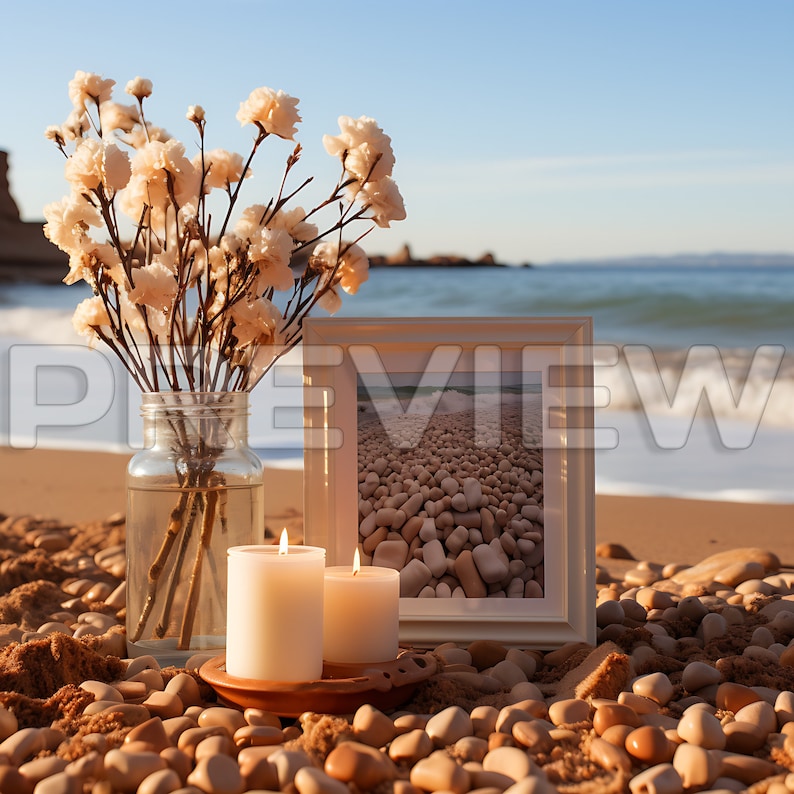May include: A white picture frame with a photo of a beach with smooth, round stones. Two white candles are in front of the frame, and a vase with white flowers is to the left. The scene is set on a beach with sand and stones.