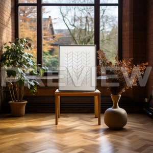 May include: A white framed print with a geometric pattern of black arrows on a white background. The print is displayed on a wooden table in front of a large window with a view of trees and a building.
