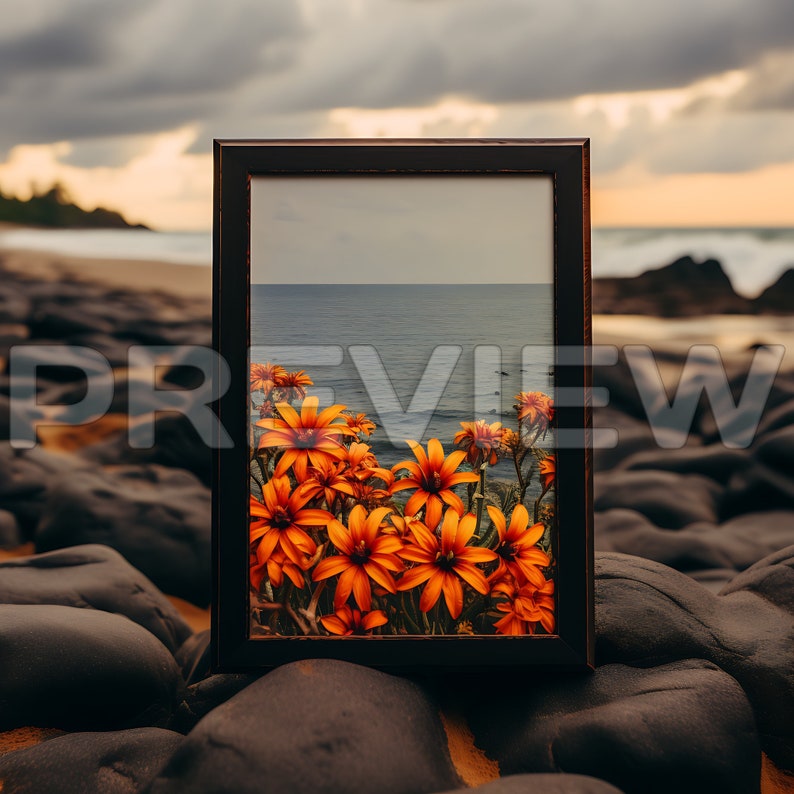 May include: A framed photograph of orange flowers in front of a blue ocean. The frame is made of dark wood and is sitting on a bed of black rocks.