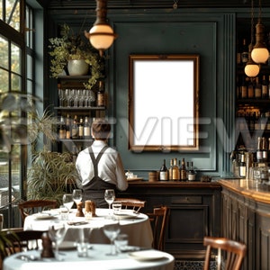 May include: A person wearing a white shirt and black overalls stands behind a bar in a restaurant. The bar is made of dark wood and has a mirror behind it. There is a large, empty picture frame hanging on the wall behind the bar. A table with white tablecloth and glasses is set in the foreground.