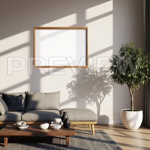 May include: A living room scene featuring a wooden-framed blank picture on a white wall. A gray sofa, ottoman, and wooden coffee table are in front of the wall. A potted plant sits to the right, and sunlight casts shadows.