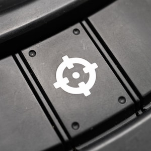 May include: Close-up of a black control panel with three rectangular buttons. The center button features a white target symbol. The buttons are slightly recessed and have a matte finish. The surrounding area is also black and textured.