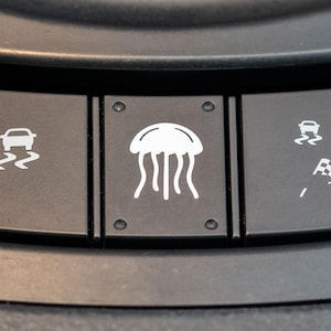 May include: Close-up of a car's dashboard with three buttons. The middle button has a white jellyfish icon, while the left and right buttons have icons depicting a car driving on a slippery surface and a checkered flag, respectively.