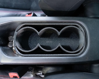 May include: A black car interior with a cup holder that has three circular openings. The cup holder is made of a textured material.