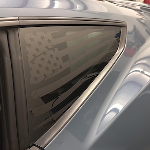 May include: A close-up of a car window with a distressed American flag design etched into the glass. The car's body is a light blue color, and the window frame is silver.