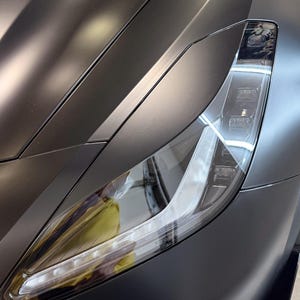 May include: Close-up of a matte black car headlight and front panel. The headlight has a clear lens with multiple LED lights. The car's body has smooth, curved lines.