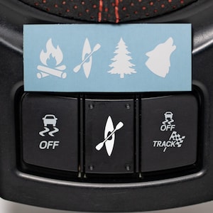 Pode incluir: Painel de controle do veículo preto com ícones brancos para recursos off-road. Um adesivo azul claro exibe símbolos de aventura ao ar livre: fogueira, caiaque, pinheiro e lobo uivando.