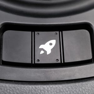 May include: Close-up of a black car dashboard button with a white rocket ship icon. The button is rectangular and has a smooth, glossy finish. The rocket icon is centered on the button.