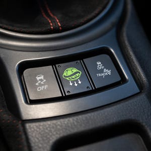 Pu&ograve; includere: Primo piano della console centrale di un'auto con tre pulsanti neri. I pulsanti sono etichettati "OFF", un logo di tartaruga verde con frecce e "OFF TRACK". La console &egrave; grigio scuro con cuciture rosse.