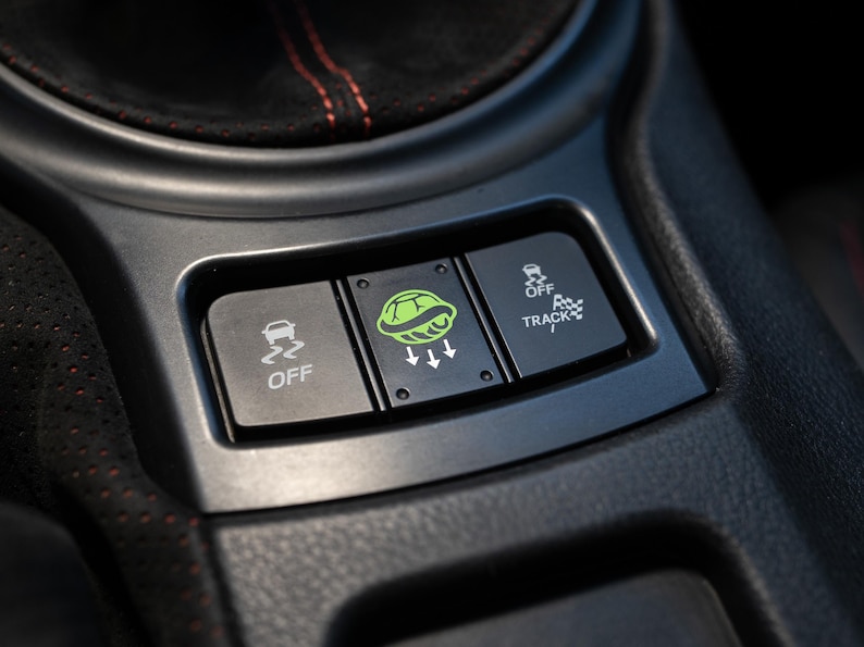 Pu&ograve; includere: Primo piano della console centrale di un'auto con tre pulsanti neri. I pulsanti sono etichettati "OFF", un logo di tartaruga verde con frecce e "OFF TRACK". La console &egrave; grigio scuro con cuciture rosse.