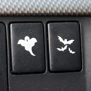 May include: Two black buttons with white Halloween-themed graphics. One button features a ghost, and the other has three bats. The buttons are set against a textured gray and black background.