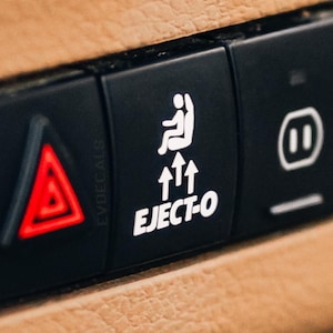 Pode incluir: Um painel de instrumentos de carro preto com um botão vermelho de luzes de emergência, um botão branco com uma figura de pau e o texto "EJECT-O" e três setas brancas apontando para cima, e um botão branco com um símbolo de pausa.