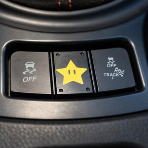 Pu&ograve; includere: Primo piano del cruscotto di un'auto con tre pulsanti neri. Il pulsante sinistro indica "OFF" con un'icona di auto che sbanda. Il pulsante centrale presenta una stella gialla con una faccia. Il pulsante destro indica "OFF TRACK" con un'icona a scacchi.