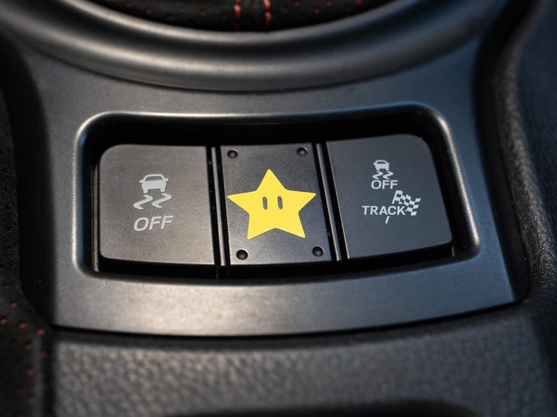 Pu&ograve; includere: Primo piano del cruscotto di un'auto con tre pulsanti neri. Il pulsante sinistro indica "OFF" con un'icona di auto che sbanda. Il pulsante centrale presenta una stella gialla con una faccia. Il pulsante destro indica "OFF TRACK" con un'icona a scacchi.