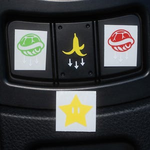 May include: Black car console with stickers featuring video game icons. Includes a green turtle shell, a yellow banana peel, a red turtle shell, and a yellow star. White arrows point downward from the icons.