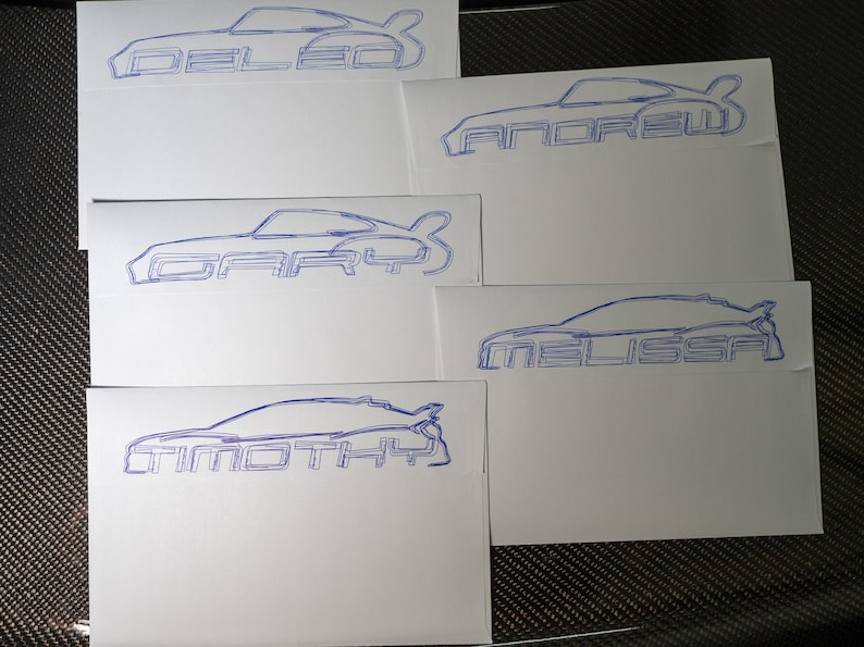 Pu&ograve; includere: Biglietti bianchi con contorni blu di auto e nomi: "DELEO", "ANDREW", "GARY", "MELISSA" e "TIMOTHY". Il design dell'auto &egrave; una coup&eacute;. I biglietti sono disposti su una superficie scura e strutturata.