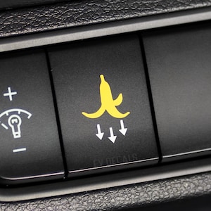 Peut inclure: Un tableau de bord de voiture noir avec un autocollant de peau de banane jaune sur un bouton. Le bouton a trois flèches blanches pointant vers le bas. Le texte "EV DECALS" est sous la peau de banane.