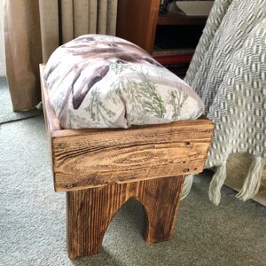 May include: A wooden footstool with a padded top featuring a floral and animal print. The footstool has a rustic brown finish and is made of reclaimed wood.