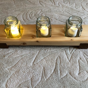 May include: A wooden candle holder with three glass jars, each containing a lit candle. The holder has a natural wood finish and is sitting on a light brown carpet.