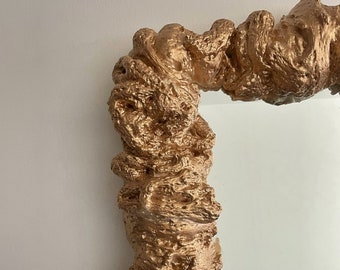 May include: A gold-colored, abstract, irregular-shaped mirror with a textured frame. The frame is made of a material that resembles a rough, organic shape.