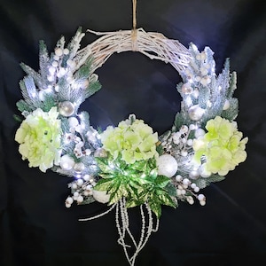 Op de afbeelding: Een krans van witte takken versierd met groene en witte bloemen, witte bessen en witte lampjes.