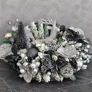 May include: A decorative centerpiece featuring a silver reindeer figurine surrounded by black and silver ornaments, flocked pine branches, dark pinecones, metallic berries, and intricate silver mesh floral accents arranged in a dense cluster.