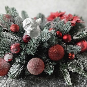 Może przedstawiać: Świąteczny stroik z ośnieżonymi gałązkami świerku, czerwonymi brokatowymi bombkami i czerwonymi kwiatami poinsecji. W centrum kompozycji znajduje się mała biała figurka aniołka, otoczona igłami sosnowymi i okrągłymi ozdobami choinkowymi.