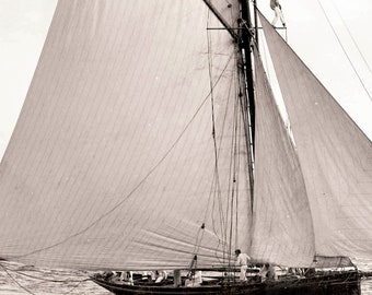 May include: A black and white photo of a sailboat with a large sail billowing in the wind. The sailboat is on the water and there are people on board.