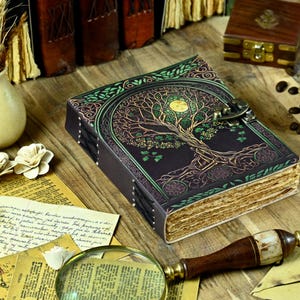 Puede incluir: Un diario de cuero marrón oscuro con un diseño de árbol de la vida en verde, dorado y morado. El diario tiene un cierre de broche y está hecho de papel reciclado.