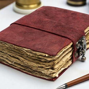May include: A rectangular journal with a deep red, textured cover and a metal latch closure. The book features thick, deckled-edge pages with a light tan, aged appearance. A brown pen with a silver tip is positioned near the bottom right corner.
