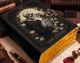 May include: A black hardcover journal featuring a tarot card illustration of two skeletons kissing surrounded by roses. The cover displays the Roman numeral VI and the text THE LOVERS. It includes a metal latch closure and gold-toned page edges.