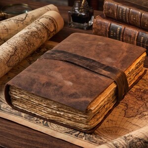 May include: A thick, rectangular journal bound in dark brown leather with a matching leather strap closure. The book features numerous pages with rough, deckled edges that appear aged and off-white in color, creating a substantial, textured appearance.