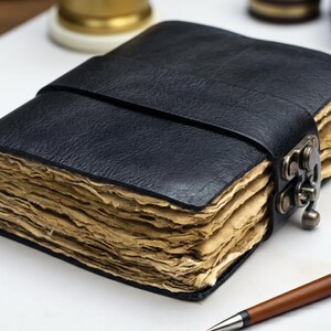 May include: A black leather-bound journal featuring a textured cover with a wide strap closure secured by a metallic latch. The journal contains thick, deckled-edge pages with a light tan, aged appearance, providing a distinct contrast to the dark cover.