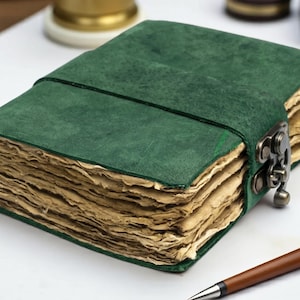 May include: A rectangular journal with a dark green suede cover and a wrap-around strap secured by a bronze-toned metal latch. The journal contains thick, deckled-edge pages with a light tan, aged appearance, visible along the side of the book.
