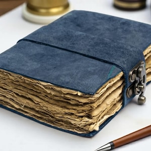 May include: A rectangular journal with a deep blue textured cover and a metal latch closure. The journal contains thick, deckled-edge pages with a light tan, aged appearance. A brown pen is positioned in the lower right corner of the frame.